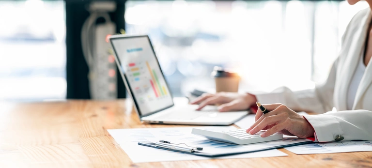 Person am Laptop mit Kugelschreiber und Taschenrechner